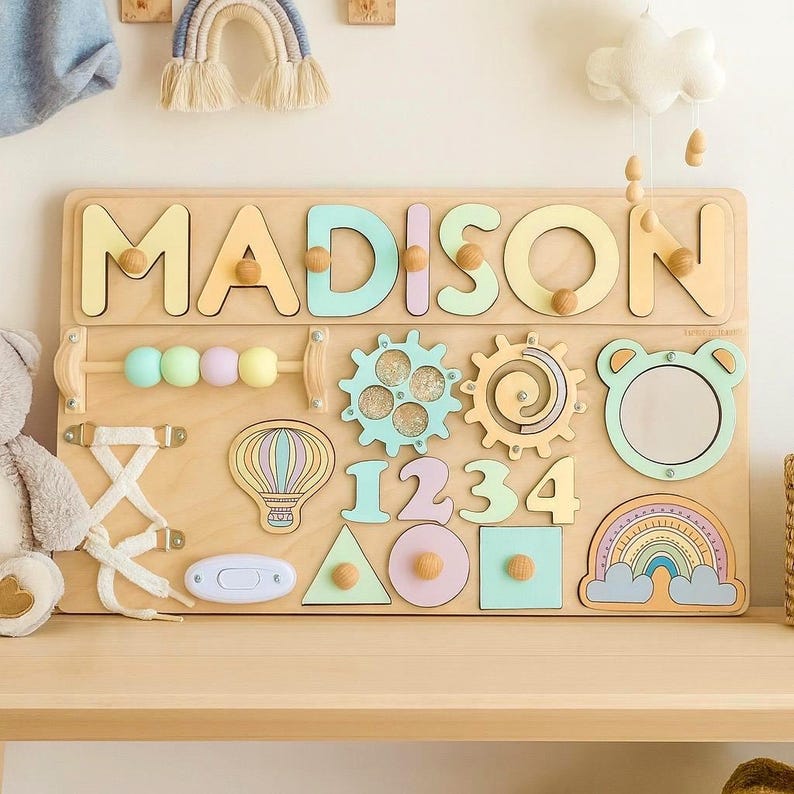 Op de afbeelding: Houten educatief speelgoed met de naam "MADISON" in pastelkleuren. Het bord bevat tandwielen, vormen, cijfers, een spiegel en een regenboogontwerp. Ontworpen voor leren en ontwikkeling in de vroege kinderjaren.