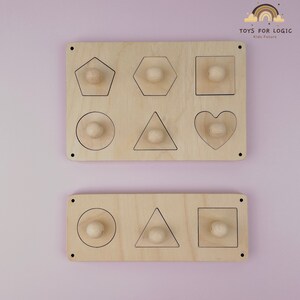 Op de afbeelding: Twee houten planken met uitsparingen in verschillende vormen. De bovenste plank heeft uitsparingen van een vijfhoek, zeshoek, vierkant, cirkel, driehoek en hart. De onderste plank heeft uitsparingen van een cirkel, driehoek en vierkant. De planken zijn gemaakt van natuurlijk hout en hebben een lichtbruine kleur. "TOYS FOR LOGIC Kids Future" is afgedrukt op de bovenste plank.