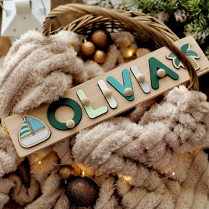 May include: A wooden name puzzle spelling "OLIVIA" in green, teal, and white letters, with a sailboat and palm tree design. The puzzle is on a wooden board with a handle, and is surrounded by a fluffy, beige blanket.