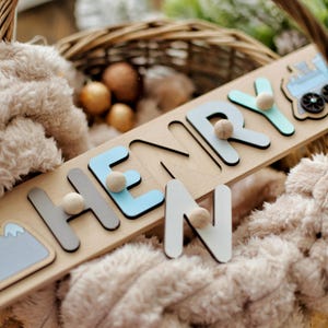 May include: A wooden name puzzle spelling "HENRY" with light blue, grey, and white letters. Each letter has a small wooden ball attached. A mountain and train graphic are on the puzzle. The puzzle is on a soft, plush surface.