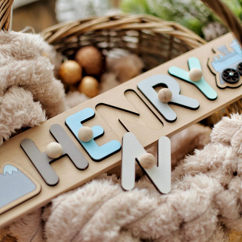 May include: A wooden name puzzle spelling "HENRY" with light blue, grey, and white letters. Each letter has a small wooden ball attached. A mountain and train graphic are on the puzzle. The puzzle is on a soft, plush surface.