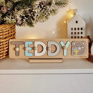 May include: A wooden personalized name puzzle with the name "TEDDY" spelled out in colorful letters. The puzzle features a cute cartoon bear holding a honey pot. The puzzle is made by Toys for Logic.