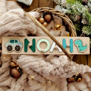 May include: A wooden name puzzle with the name "NOAH" spelled out in colorful letters. The puzzle pieces are shaped like a car, a dinosaur, and the letters of the name. The puzzle is on a wooden background.