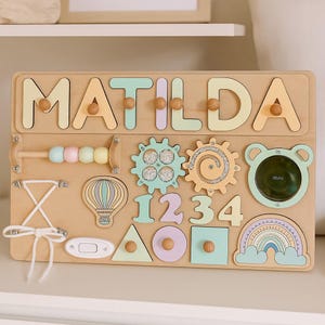 Op de afbeelding: Houten activiteitenbord met de naam "MATILDA" in pastelkleuren. Voorzien van tandwielen, cijfers, vormen, een regenboog en een luchtballon. Inclusief een kralenpuzzel en een strik. Een leuk en leerzaam speelgoed.
