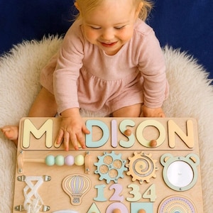 Op de afbeelding: Houten educatief speelgoed met de naam "MADISON" in pastelkleurige letters. Het bord bevat verschillende vormen, tandwielen, cijfers en een spiegel, ontworpen voor vroeg leren en sensorische exploratie. Het speelgoed ligt op een witte, zachte ondergrond.