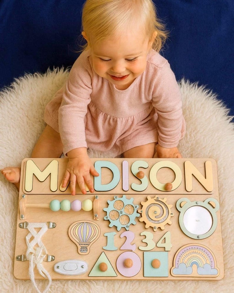 Op de afbeelding: Houten educatief speelgoed met de naam "MADISON" in pastelkleurige letters. Het bord bevat verschillende vormen, tandwielen, cijfers en een spiegel, ontworpen voor vroeg leren en sensorische exploratie. Het speelgoed ligt op een witte, zachte ondergrond.