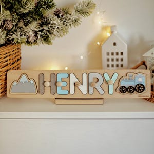 May include: Personalized wooden puzzle with the name "HENRY" in blue, white, and gray letters. The puzzle features a mountain and a blue train with black wheels.