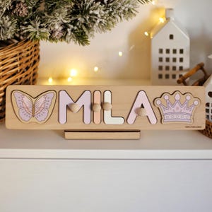 May include: Wooden name puzzle spelling "MILA" with a butterfly and crown design. The letters are in pastel colours, with wooden pegs for easy handling. The puzzle is designed for children.