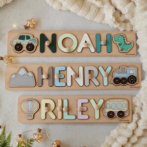 May include: Three wooden personalized name puzzles with colorful letters and illustrations. The top puzzle has a green monster truck, the middle puzzle has a gray mountain, and the bottom puzzle has a hot air balloon. Each puzzle has a small wooden knob at the end of each letter.