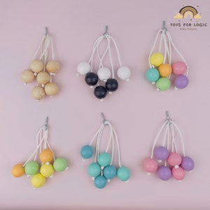 Peut inclure: Six ensembles de boules en bois suspendues à des cordons blancs. Chaque ensemble comprend cinq boules de couleurs différentes : bois naturel, noir et blanc, jaune, vert et bleu, et violet et rose. Les boules sont attachées aux cordons à l’aide d’un crochet argenté. L’arrière-plan est rose pâle.