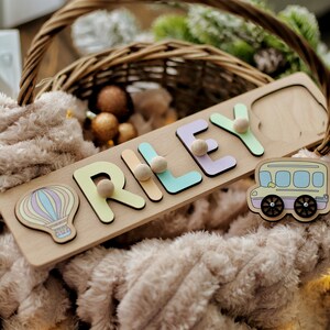 May include: A wooden name puzzle with the name "RILEY" spelled out in colorful letters. The puzzle pieces are removable and have small wooden knobs for easy handling. The puzzle also includes a hot air balloon and a school bus.