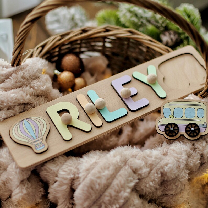 May include: A wooden name puzzle with the name "RILEY" spelled out in colorful letters. The puzzle pieces are removable and have small wooden knobs for easy handling. The puzzle also includes a hot air balloon and a school bus.