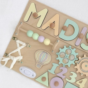 Op de afbeelding: Houten activiteitenbord met de naam "MADISON" in pastelkleuren. Voorzien van een kralenlabirint, tandwielen, vormen, een lichtschakelaar en een illustratie van een luchtballon. Een leuk en leerzaam speelgoed voor peuters.