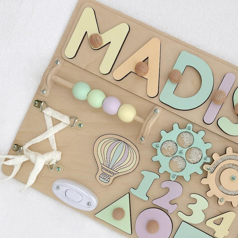 Op de afbeelding: Houten activiteitenbord met de naam "MADISON" in pastelkleuren. Voorzien van een kralenlabirint, tandwielen, vormen, een lichtschakelaar en een illustratie van een luchtballon. Een leuk en leerzaam speelgoed voor peuters.