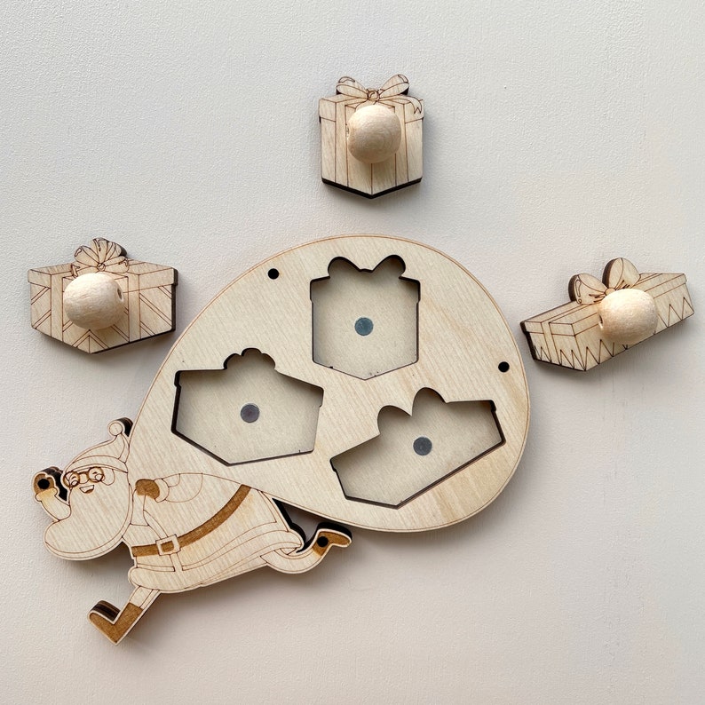 Puede incluir: Puzzle de madera con Santa Claus llevando una bolsa de regalos. El rompecabezas tiene cuatro piezas: Santa, una bolsa y tres regalos. Los regalos tienen un pomo en la parte superior para facilitar su manejo.