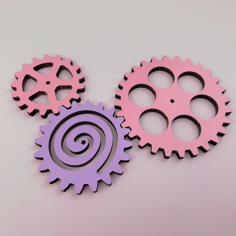 May include: Three wooden gears, one pink with six teeth and five holes, one pink with eight teeth and a spiral design, and one purple with a spiral design.