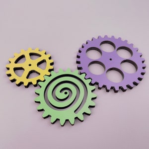 May include: Three wooden gears interlocked. The largest gear is purple with six teeth and a spiral design. The middle gear is green with a black spiral design. The smallest gear is yellow with six teeth.