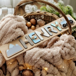 May include: Wooden name puzzle spelling "HENRY" with blue, gray, and white letters, a mountain, and a train. The puzzle is on a light wood base. The background includes a fluffy blanket and a wicker basket.