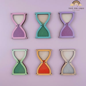 Könnte beinhalten: Sechs Holz-Sanduhren mit unterschiedlich farbigem Sand. Die Sanduhren sind an einer rosa Wand befestigt. Die Oberseite jeder Sanduhr ist in einer anderen Farbe gehalten: rosa, blau, lila, grün, orange und naturbelassenes Holz. Der Sand in jeder Sanduhr ist in einer anderen Farbe: lila, grün, lila, grün, orange und weiß.