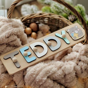 May include: A wooden puzzle spelling "TEDDY" with grey, blue, and white letters, featuring a bear illustration. The puzzle includes wooden knobs for easy handling. The puzzle is set in a wicker basket with a plush toy.