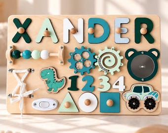 May include: A rectangular wooden busy board featuring the name XANDER in various shades of green and blue. It includes a bead abacus, two spinning gears, a bear-shaped mirror, a dinosaur cutout, numbers 1234, a white lace-up, and a toy truck.