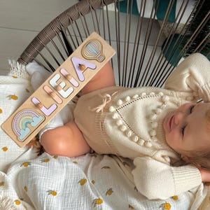May include: A wooden name puzzle spelling "LEIA" with a rainbow and hot air balloon design. The puzzle is light brown with pastel-coloured letters and illustrations. The puzzle is resting on a white blanket with a yellow lemon pattern.