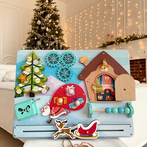 Puede incluir: Un tablero de actividades de madera con tem&aacute;tica navide&ntilde;a con una variedad de actividades para ni&ntilde;os. El tablero presenta una casa con una puerta que se abre, un &aacute;rbol de Navidad con una estrella giratoria, una figura de Pap&aacute; Noel, un reno tirando de un trineo y un laberinto de cuentas.