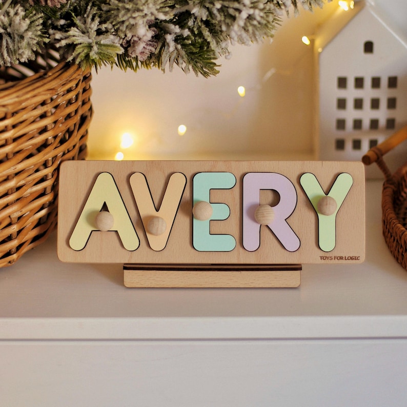 May include: A wooden puzzle with the name AVERY spelled out in pastel colors. Each letter is a separate piece with a wooden knob on the back. The puzzle is on a wooden stand with the words "TOYS FOR LOGIC" printed on the bottom.