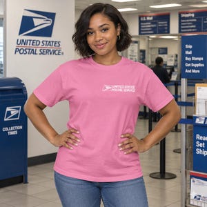 Puede incluir: Una camiseta rosa de manga corta con el logotipo del United States Postal Service impreso en blanco en el pecho izquierdo. La prenda tiene cuello redondo y un corte estándar, confeccionada en tela de color sólido con un diseño sencillo.