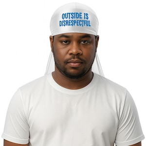 Può includere: Un durag bianco con la scritta "OUTSIDE IS DISRESPECTFUL" in blu. Il durag è realizzato in un materiale setoso e viene indossato sulla testa. La persona nell'immagine indossa una maglietta bianca.