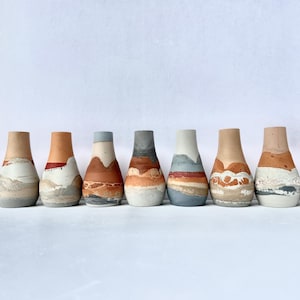 May include: Ten small ceramic vases with a layered, abstract design in shades of brown, grey, and white. The vases are arranged in a row on a white surface.