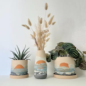 May include: Three ceramic planters and a vase with a sunset design. The planters and vase are beige with a painted orange sun, blue and gray water, and white clouds. The vase holds dried beige grasses. The planters hold a succulent and a leafy plant.