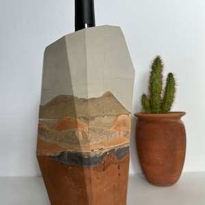 May include: A concrete wine bottle holder with a geometric design. The holder is painted in shades of brown, gray, and black, resembling a desert landscape.