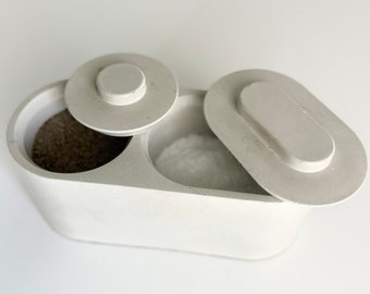 May include: White ceramic salt and pepper shaker set with lids. The shakers are oval shaped and have a separate compartment for each spice. The lids are round and have a small knob on top.