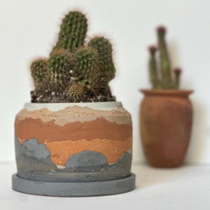 Puede incluir: Una planta de cactus verde en una maceta de cerámica en capas, texturizada, gris, naranja y blanca con un platillo gris a juego. Otro cactus en una maceta de terracota está en el fondo. Las macetas están sobre una superficie blanca.