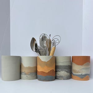 Op de afbeelding: Vijf betonnen bestekhouders met een gelaagd, abstract ontwerp. De houders zijn in tinten grijs, bruin en oranje. De houders zijn in een rij van links naar rechts gerangschikt.