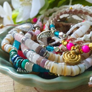 May include: A collection of colorful beaded bracelets in a light green bowl. The bracelets feature various beads in shades of pink, blue, white, and gold. Some bracelets have charms, and the bowl sits on a wooden surface.