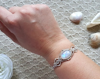 Könnte beinhalten: Ein weißes Makramee-Armband mit einem großen ovalen Mondstein. Das Armband wird am Handgelenk einer Person getragen und der Hintergrund ist ein beigefarbener Stoff mit Muscheln.