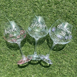 May include: Three clear gin glasses with names and floral patterns. The names "Nicola", "Jamie", and "Christina 30" are written in cursive. Each glass features a different colour floral design around the base, set against a green background.