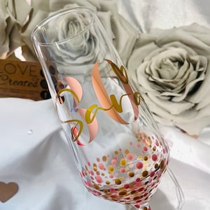 May include: A clear glass champagne flute with a gold and pink polka dot pattern. The flute has a gold vinyl sticker with the name "Sara" and the number "30" in a rose gold vinyl sticker.