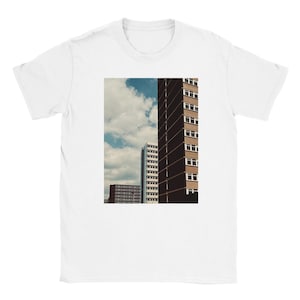 Puede incluir: Camiseta blanca con una imagen cuadrada de edificios altos contra un cielo nublado. Los edificios son marrones, blancos y grises, con muchas ventanas. El cielo es azul con nubes blancas. La camiseta es de cuello redondo.