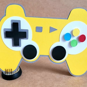 May include: A yellow cardboard cutout of a video game controller with black buttons, a grey directional pad, and colourful buttons.