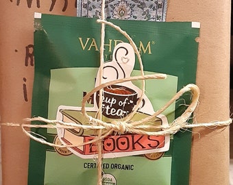 May include: A brown paper package wrapped with twine and a blue patterned ribbon. The package contains a book with the words "Fiction" and "Mystery" written on the front. There is also a green tea bag with a sticker that says "A cup of tea & books" and a white heart.