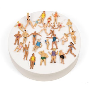 May include: Miniature figurines of people in swimwear and beach attire are arranged on a white, circular base. The figures are in various poses, some holding beach balls, and are in a variety of colours. The base is a circular shape.