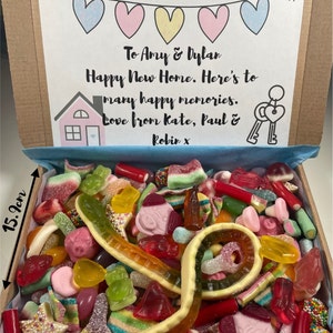 May include: A box of sweets, 21.8 cm wide and 15.9 cm high, overflowing with a variety of colorful candies. The box lid features a banner of hearts and a message: "To Amy & Dylan, Happy New Home. Here's to many happy memories. Love from Kate, Paul & Robin x."