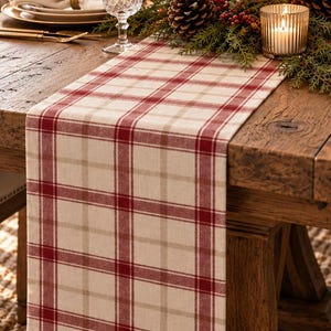 Puede incluir: Una mesa de comedor de madera rústica con un camino de mesa a cuadros en crema y rojo. La mesa está puesta con platos blancos, servilletas dobladas, cubiertos dorados y cristalería transparente. Centro de mesa con piñas, vegetación y velas blancas.