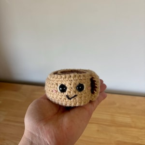 Puede incluir: Una taza de café de ganchillo con una cara sonriente. La taza es de color beige con un interior marrón y un asa pequeña. Los ojos son negros y las mejillas rosas. La taza se sostiene en una mano.
