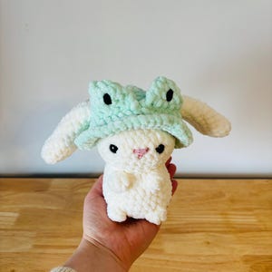 Puede incluir: Conejo de crochet blanco con un gorro de rana de crochet verde con ojos negros.