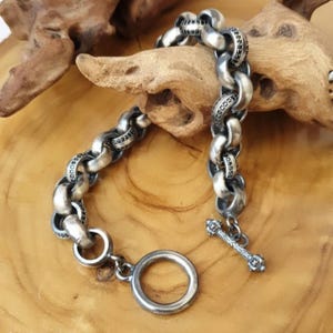 Peut inclure: Un bracelet en argent avec une chaîne épaisse et un fermoir à glissière. La chaîne a un design texturé avec de petites touches noires.