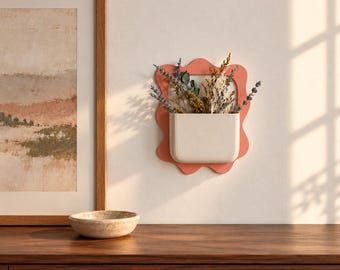 May include: A wall-mounted planter featuring a wavy, coral-colored frame and a solid off-white pocket container. The planter holds a collection of dried flowers and stems, including lavender and various grasses, arranged in a vertical display.